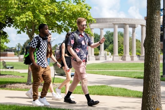 Three young students walk across a green campus in front of a columned scultpure