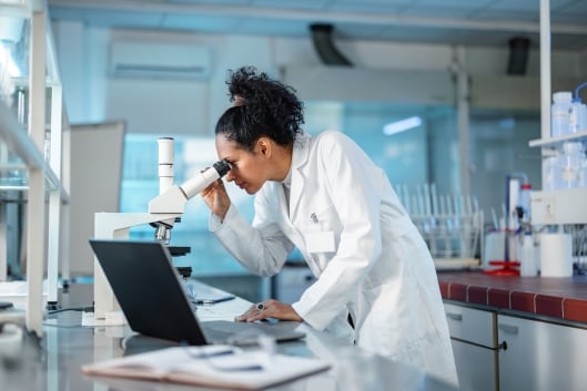 Young Hispanic scientist wearing a lab coat, looking under microscope while using laptop in a laboratory