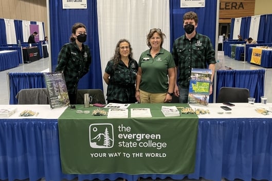 Four people stand behind a table with the words "the Evergreen State College" on a banner