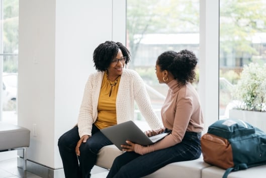 A stock photo of a Black woman professor speaking to an engaged student holding a laptop. The professor is smiling and the two appear to be in a good conversation in front of a wall of windows.