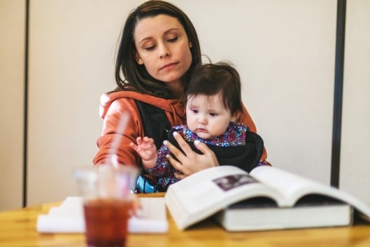 A young mother sitting at a table at the library balances a child on her lap as she takes notes on an open notebook next to two textbooks (one open, one closed). 