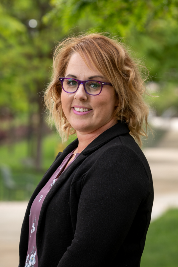 Jessica Oyler, a light-skinned woman with reddish-brown wavy hair, smiles for a head shot wearing a black blazer, purple shirt and purple glasses.