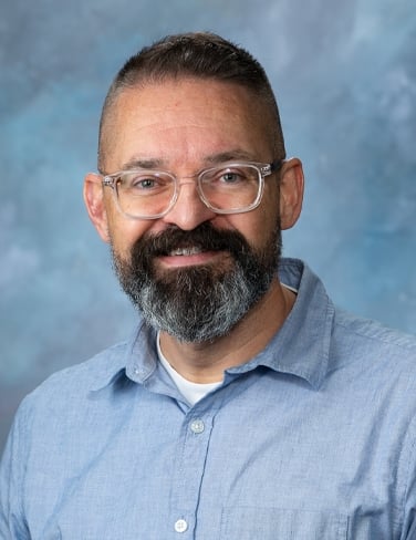 Christopher Waugh smiles for a head shot in a blue buttoned shirt against a blue background.