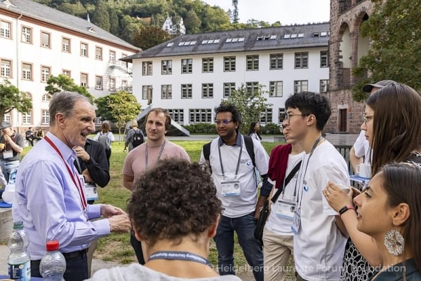 An older, white man talks with young students of various nationalities. 