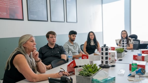 Jane Swift, a light-skinned woman with long gray hair, sits at a conference room table with several students participating in Education at Work experiences.