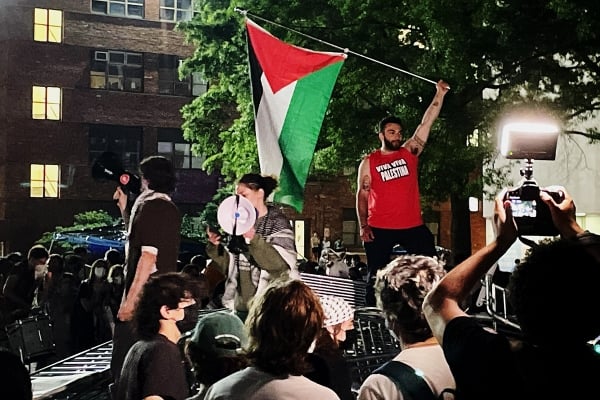 A photo of a protester waving a Palestinian flag atop a stack of barricades.