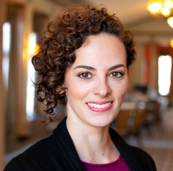 Sara Abelson, a light-skinned woman with curly dark hair, smiles for a headshot in a black blazer and purple top.