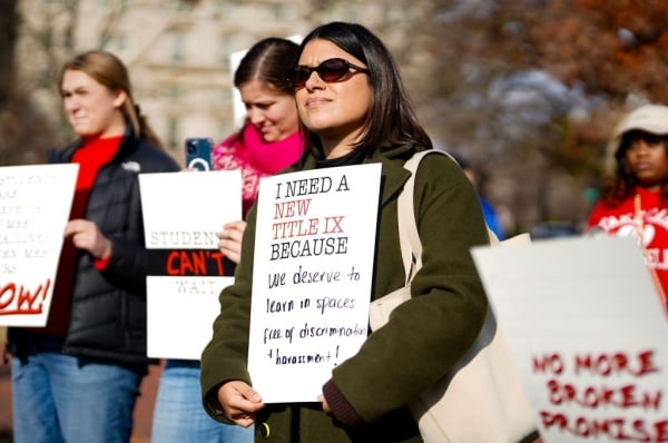 A person holds a white sign that says "I need a new Title IX because we deserve to learn in spaces free of discrimination and harassment"