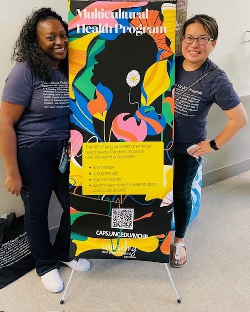 Two counseling staff members wear matching gray t-shirts and stand next to a banner advertising services for the Multicultural Health Program at UNC Chapel Hill.