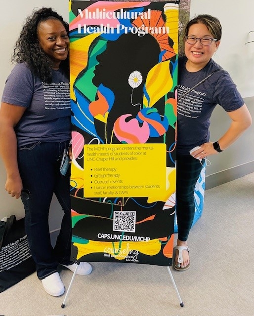 Two counseling staff members wear matching gray t-shirts and stand next to a banner advertising services for the Multicultural Health Program at UNC Chapel Hill.