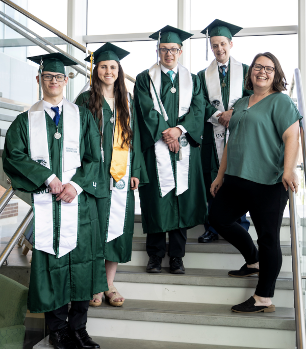 Four students pose in green graduation regalia on a flight of stairs accompanied by a mature woman in a green blouse.