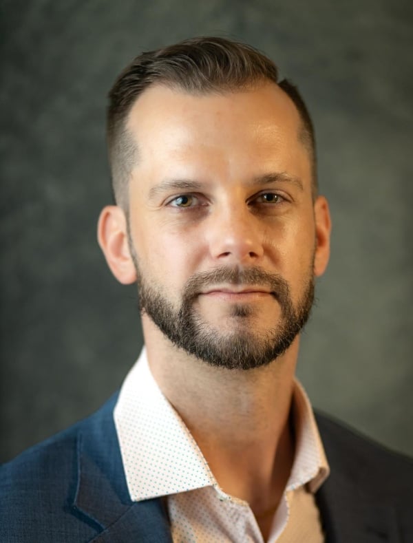 Michael Crook poses for a headshot on a gray background wearing a light shirt and dark coat.