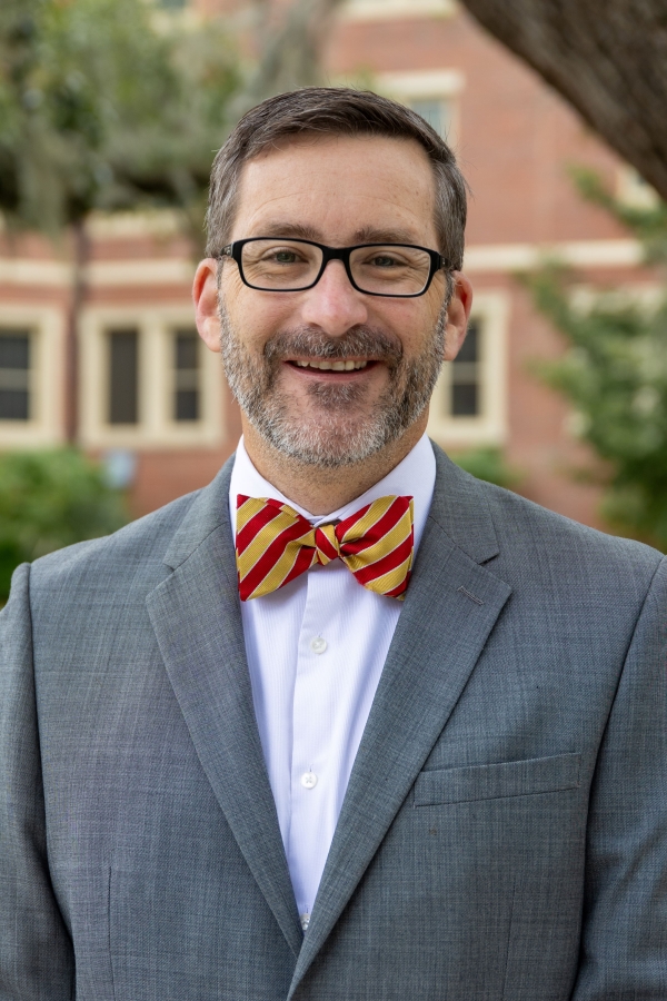 Rob Liddell smiles for a headshot wearing black framed glasses, a red and yellow striped bowtie and a gray suit coat