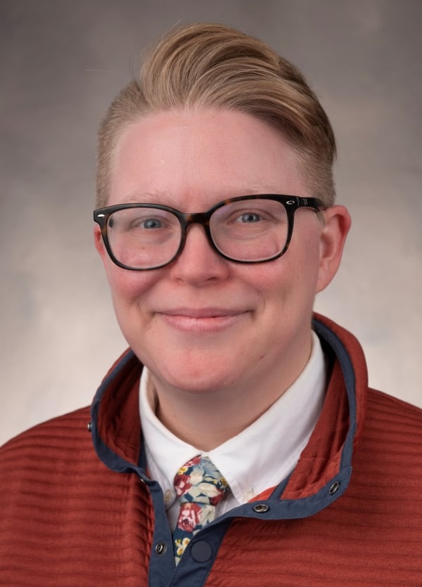 Emily Krechel smiles for a headshot wearing a red polo, multicolor tie and black frame glasses