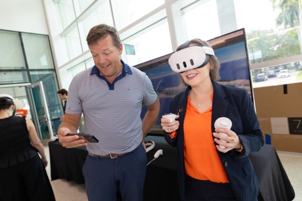 A man in a blue polo shirt holds his phone while a woman in a blue blazer and orange blouse wears a VR headset and holds two controllers.