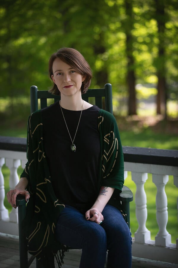 A photo of Courtney Floyd sitting in a green wooden chair, wearing jeans and a green top with a long pendant necklace.