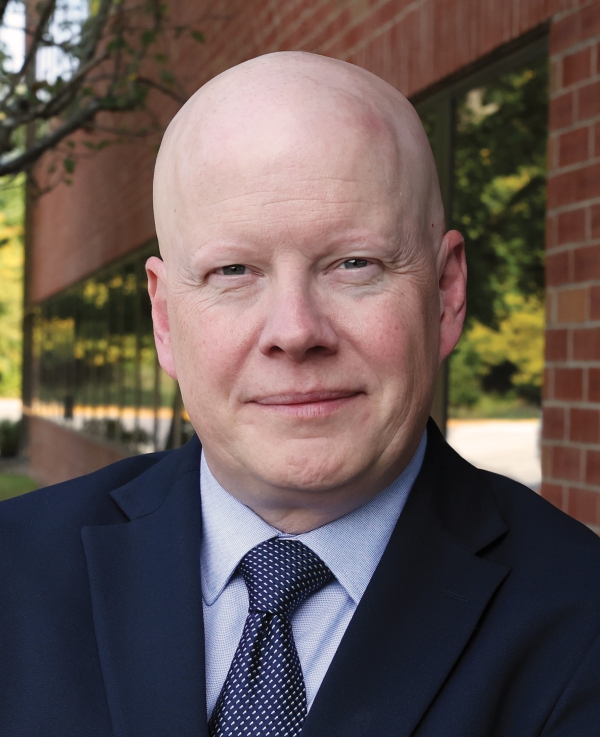 A light-skinned bald man wearing a blue suit, light blue shirt, and blue and white patterned necktie.