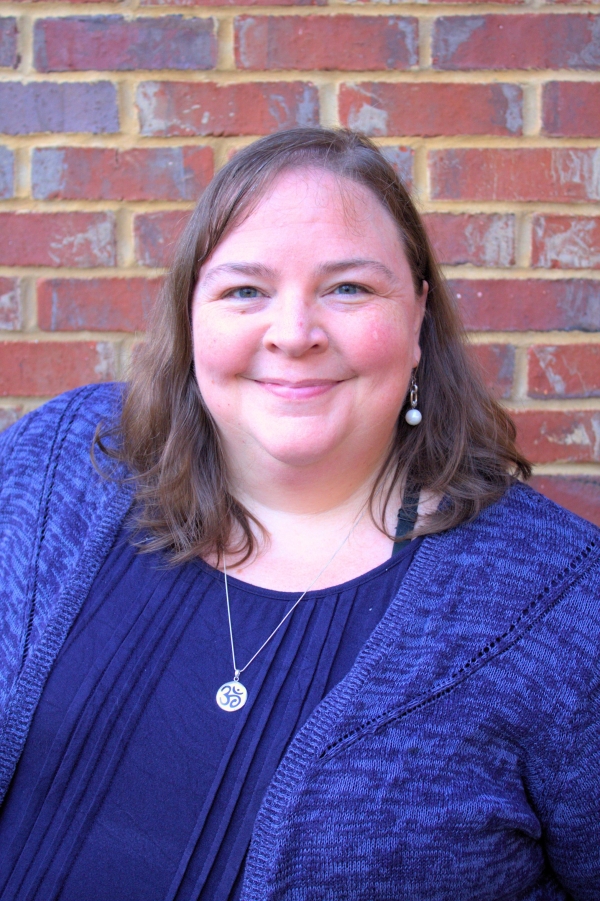 Liz Norell, a light-skinned woman with shoulder-length brown hair, wearing a purple-blue sweater set and a silver pendant necklace.