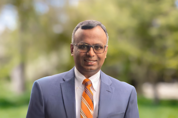 Mallik Sundharam, a man with light brown skin and graying hair, wearing glasses and a blue suit with an orange striped tie.