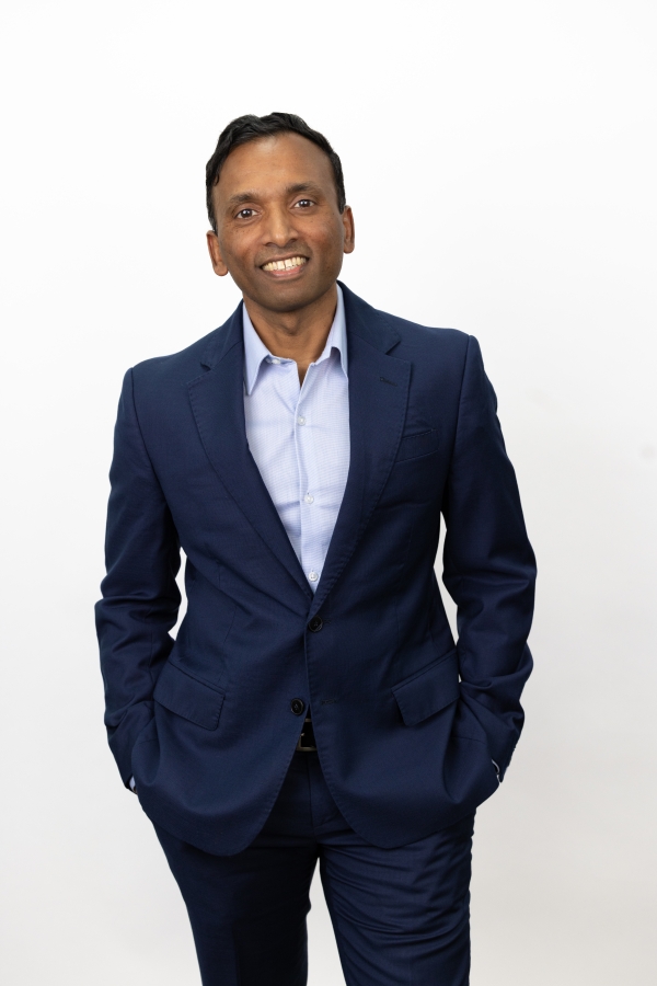 Shravan Goli, a brown-skinned man wearing a collared shirt and jacket but no tie.