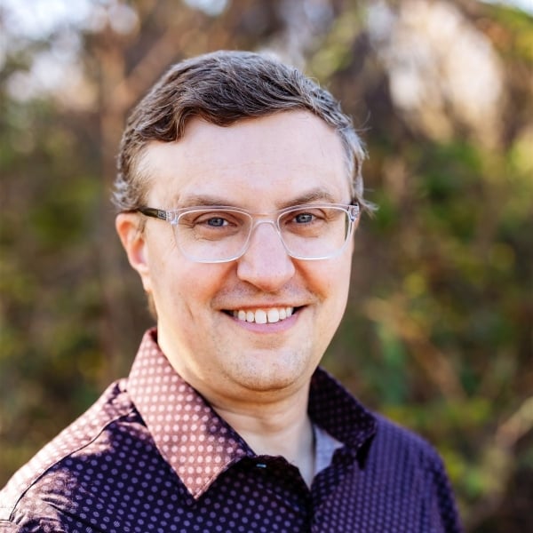 A light-skinned man wearing clear-framed glasses and a shirt with a dot pattern.