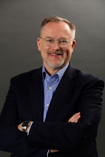 Scott Jeffe, a light-skinned man wearing wire-frame glasses and a dark blazer over a blue-and-white checkered collared shirt.