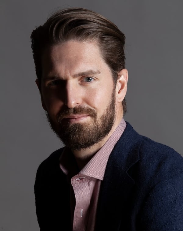 A light-skinned man with brown hair and a beard, wearing a collared shirt and a blue blazer.