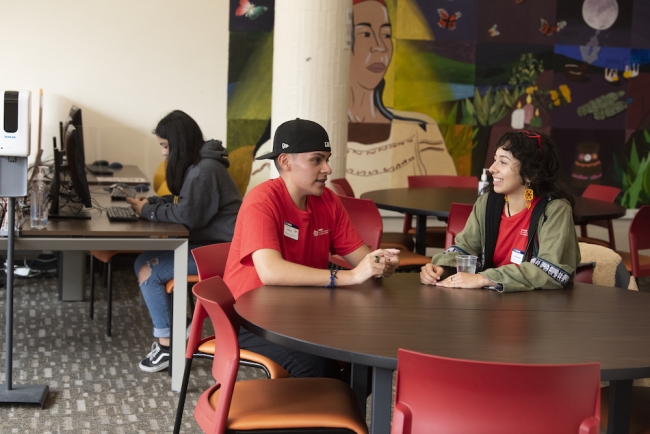 Three students work and chat in UC Davis's Center for Chicanx and Latinx Academic Student Success with a colorful mural in the background.