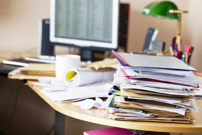 Desk rather in disarray, full of papers, pens, a coffee mug, lamp, computer and other items