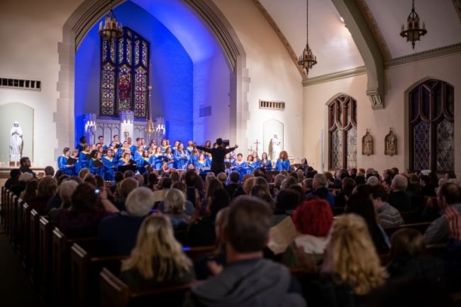 A group of students sits in the high-ceilinged chapel at Notre Dame College watching a choir in blue and gold robes.