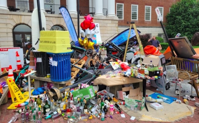 A colorful array of trash, bottles, stolen signs, electric scooters, trash cans, a lamppost and more are scattered around a statue of the University of Maryland's terrapin mascot.