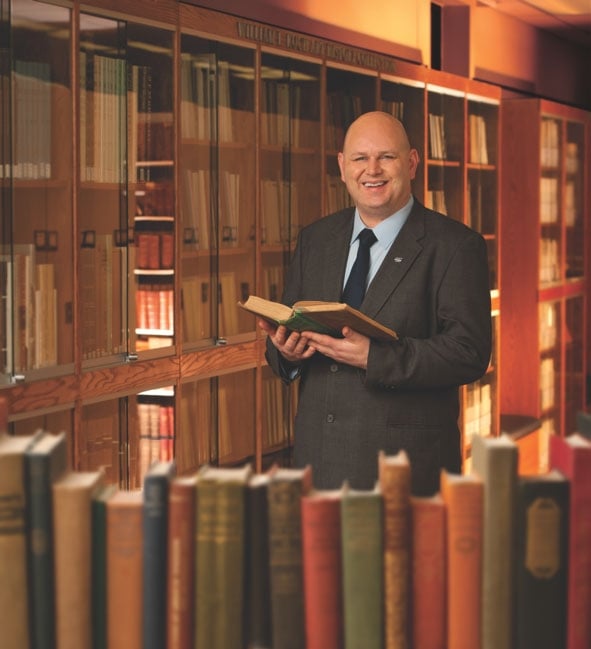 Alan Bearman, a bald man with light skin wearing a business suit, smiles holding a book in a library.