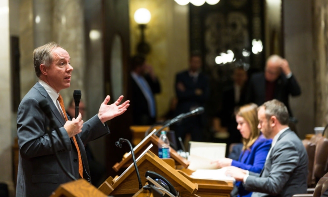 A man in a suit and tie speaks at a podium to a room of legislators