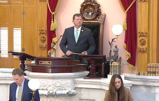 A man in a suit stands at a raised podium above another man and a woman, backed by gold and red curtains.