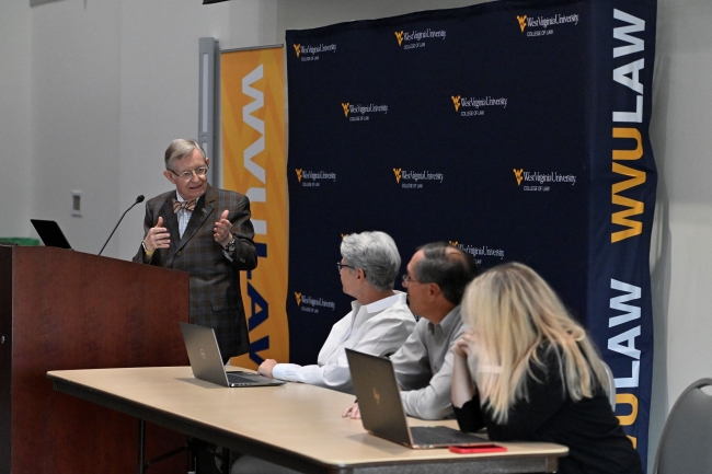 A man in a bow tie and glasses at a podium turns to three panelists