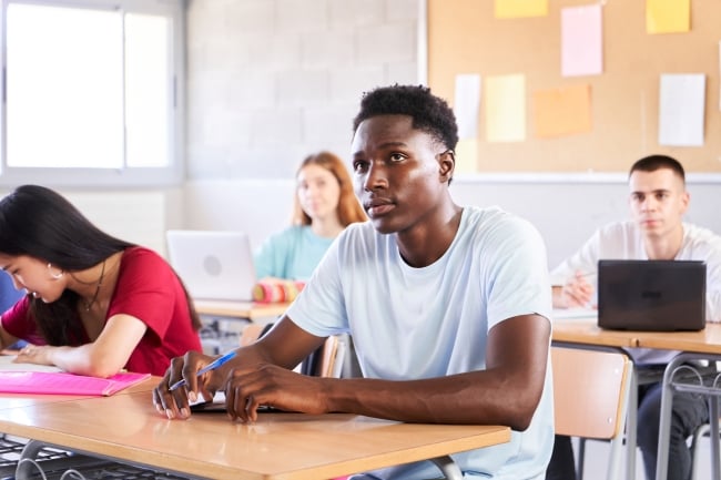 A Black male student sits at a desk with peers sitting next to and beside him