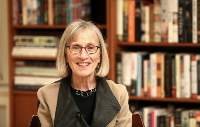 A photo of Claudia Goldin, a light-skinned woman with shoulder-length light hair, pictured in front of a bookcase.