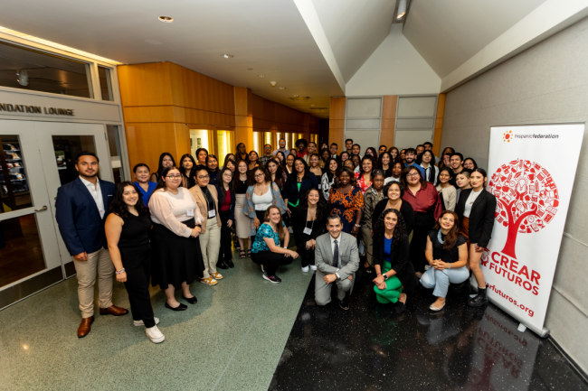 A group of Latino mentors smile for a photo in front of a banner with a red tree logo and CREAR Futuros beneath it.