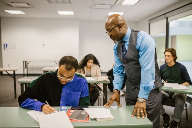 A professor looks over the shoulder of a student engaged in a classroom, with other students working at desks in the background.