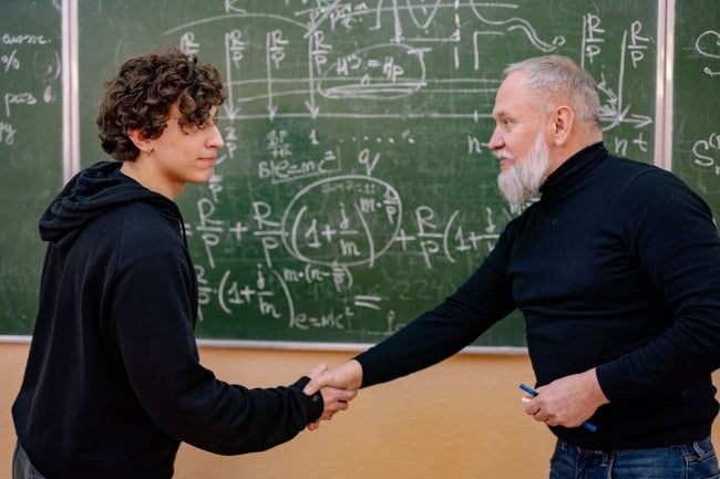 A professor standing in front of a chalkboard shakes a student's hand.