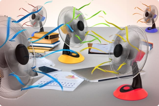 Five fans of different colors blow breeze around a desk cluttered with school supplies, laptops, papers and books.