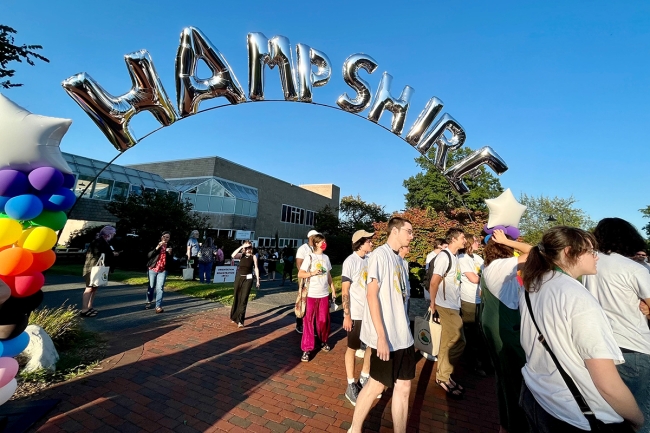 Students in white shirts walk beneath a large balloon arc, reading "Hampshire" in silver letters.