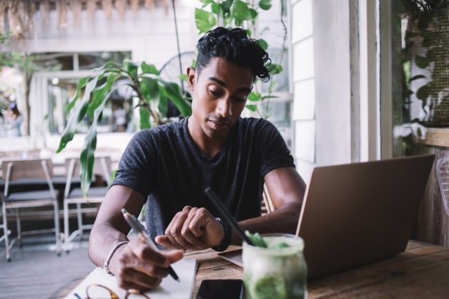 Young man sitting on a terrace or at a cafe looks at his watch as he's writing on a notepad with his laptop open.