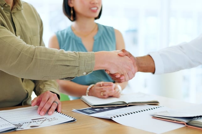 Two people shake hands in front of a young woman at a table with books and papers 