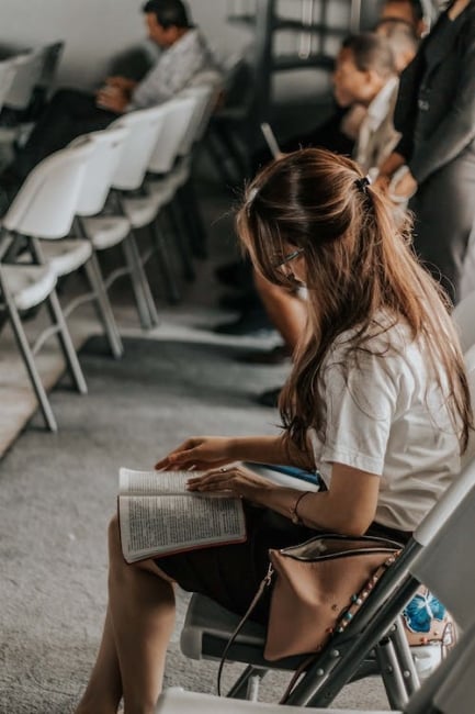 Close-up of a student sitting in a room reading a book with lines of chairs and a few people of different ages sitting and waiting