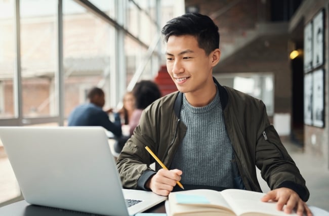 A young man works with his laptop open using a pencil and notebook, studying in a common area during the daytime.