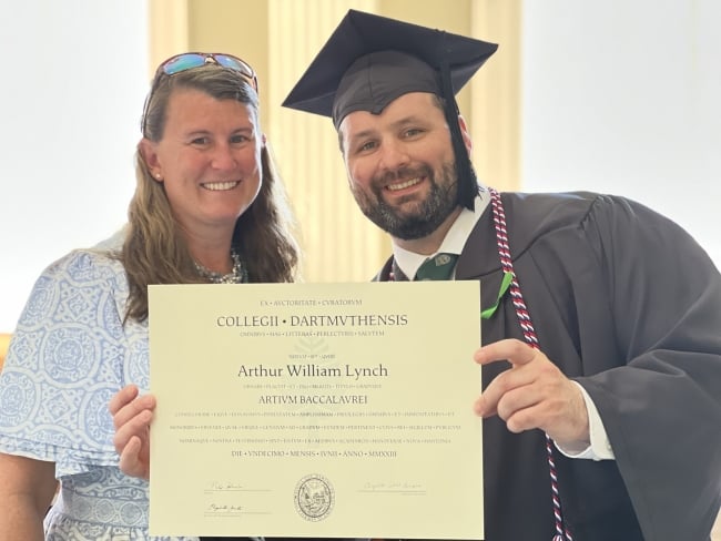 Photo of a light-skinned woman and a light-skinned man in academic regalia smiling while holding a diploma.