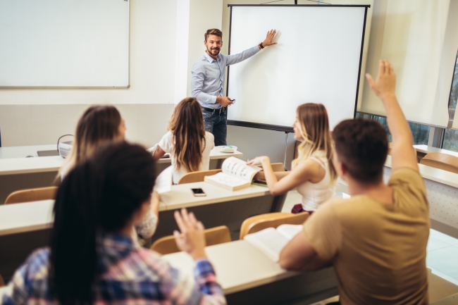 Smiling teacher standing in front of students and showing something on whiteboard in classroom.