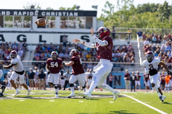 A football player in a red jersey and helmet throws a football across a field of other players as a crowd looks on