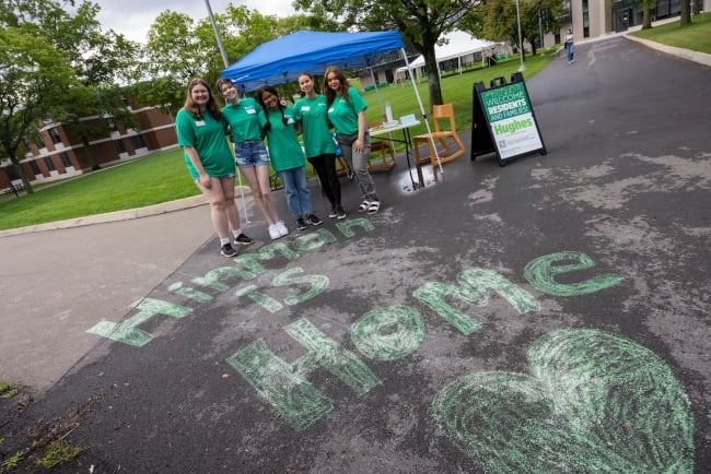 Residential life student staff members at Binghamton University wearing green T-shirts and standing in front of a chalk drawing reading "Hinman Is Home" greet first-year students.